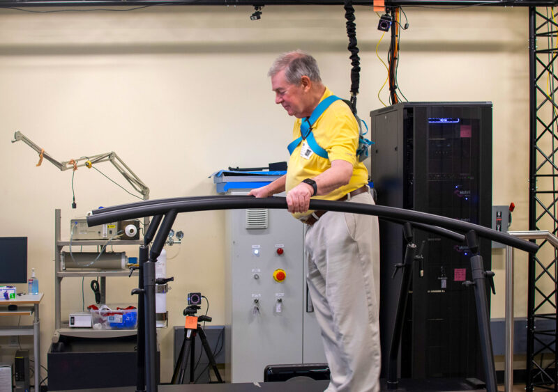 An older man in a harness walks on a treadmill in a laboratory, holding onto black rails, with scientific equipment and computers in the background.