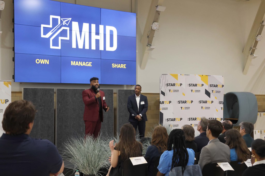 Two presenters stand on stage in front of an audience, with a large screen displaying "MHD" and a banner reading "START UP" in the background.