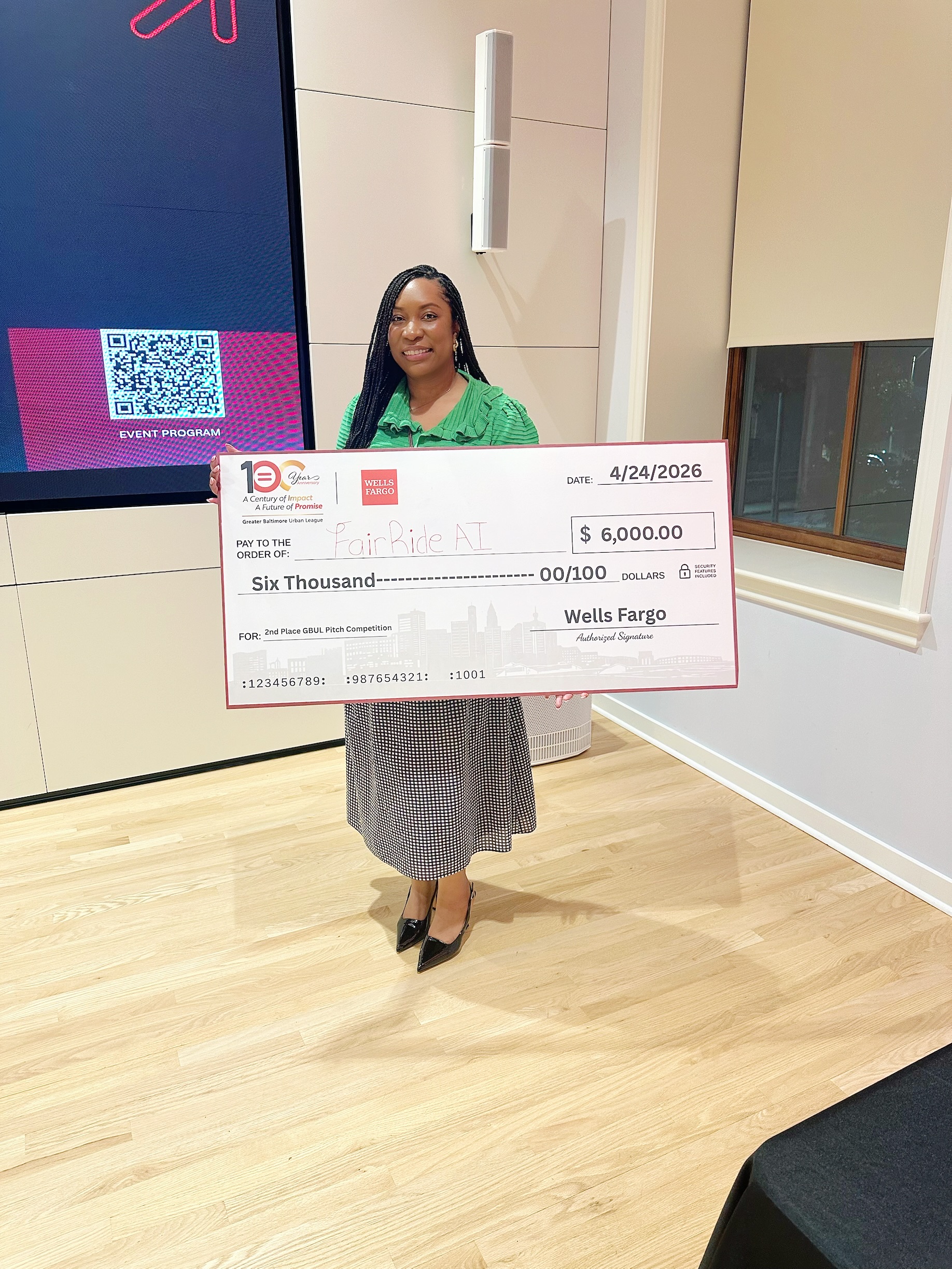 A woman stands indoors holding a large check for $6,000 made out to FairRide ATL from Wells Fargo, dated 4/24/2026.
