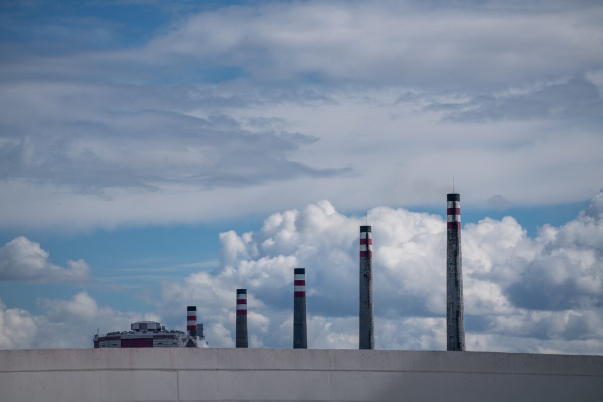 Six tall industrial chimneys with red and white markings rise against a cloudy blue sky; a large building is partially visible on the left.