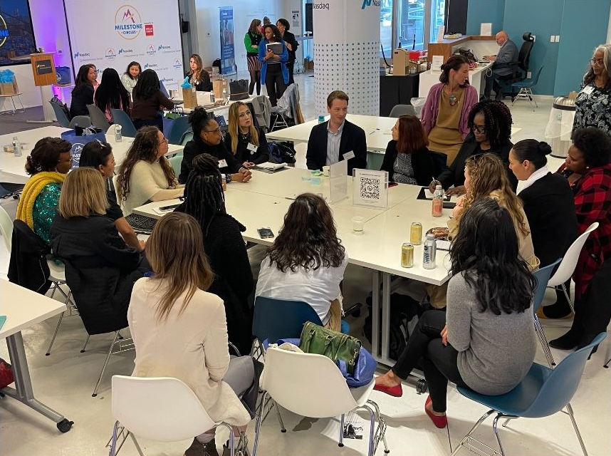 A group of people sit around a table in discussion at an indoor event, with others gathered at tables in the background.