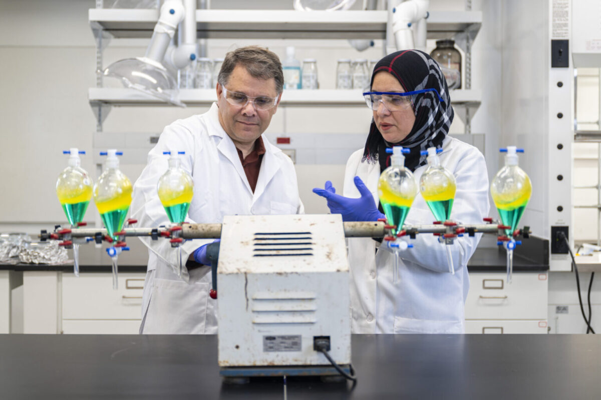 Two scientists in lab coats and safety goggles examine equipment with six flasks containing green and yellow liquids in a laboratory setting.