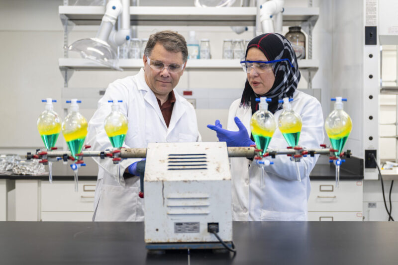 Two scientists in lab coats and safety goggles examine equipment with six flasks containing green and yellow liquids in a laboratory setting.