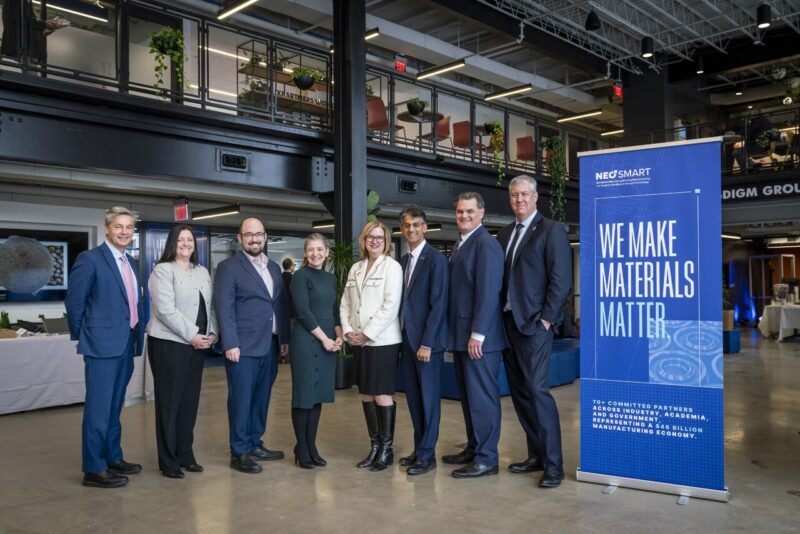 Eight people in business attire stand in a row, posing for a group photo indoors next to a blue "We Make Materials Matter" banner. Modern office space and plants are visible in the background.