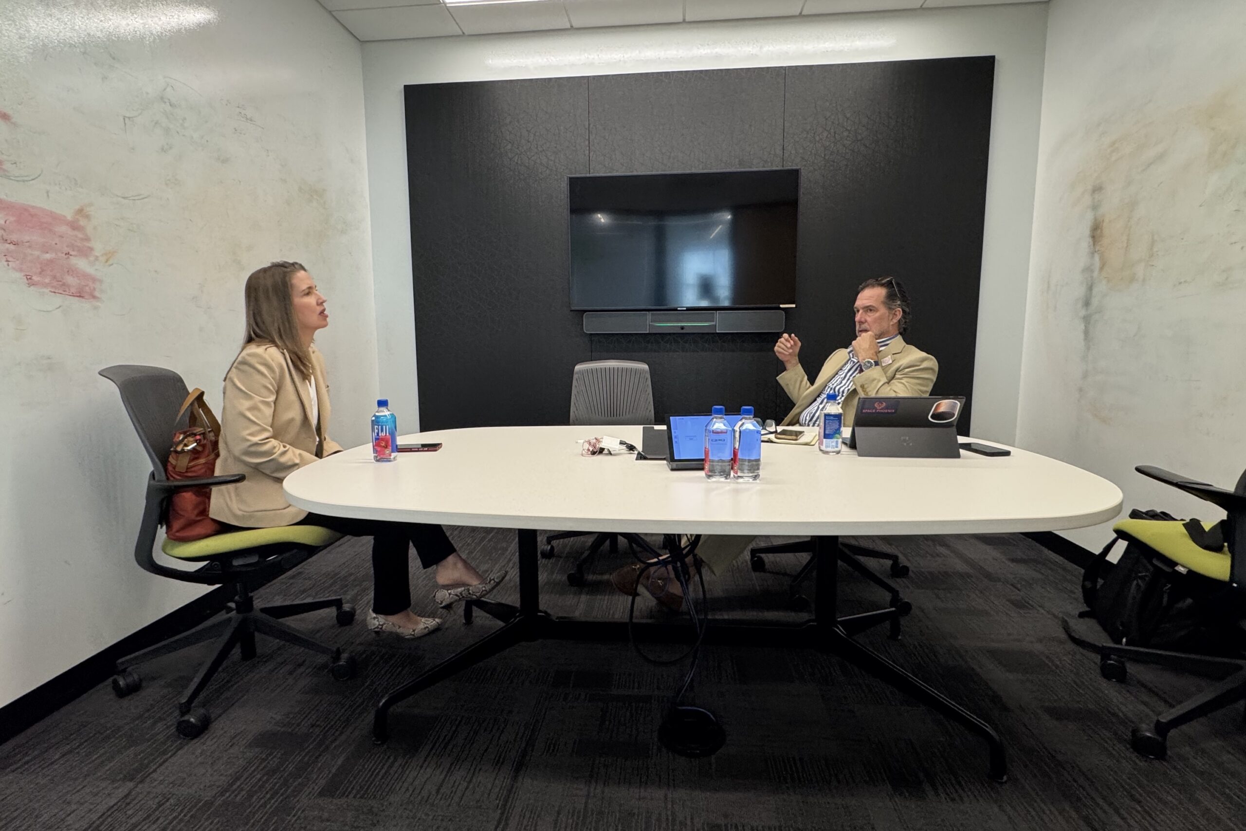 Two people in business attire sit across from each other at a conference table with laptops, papers, and water bottles in a modern office meeting room.