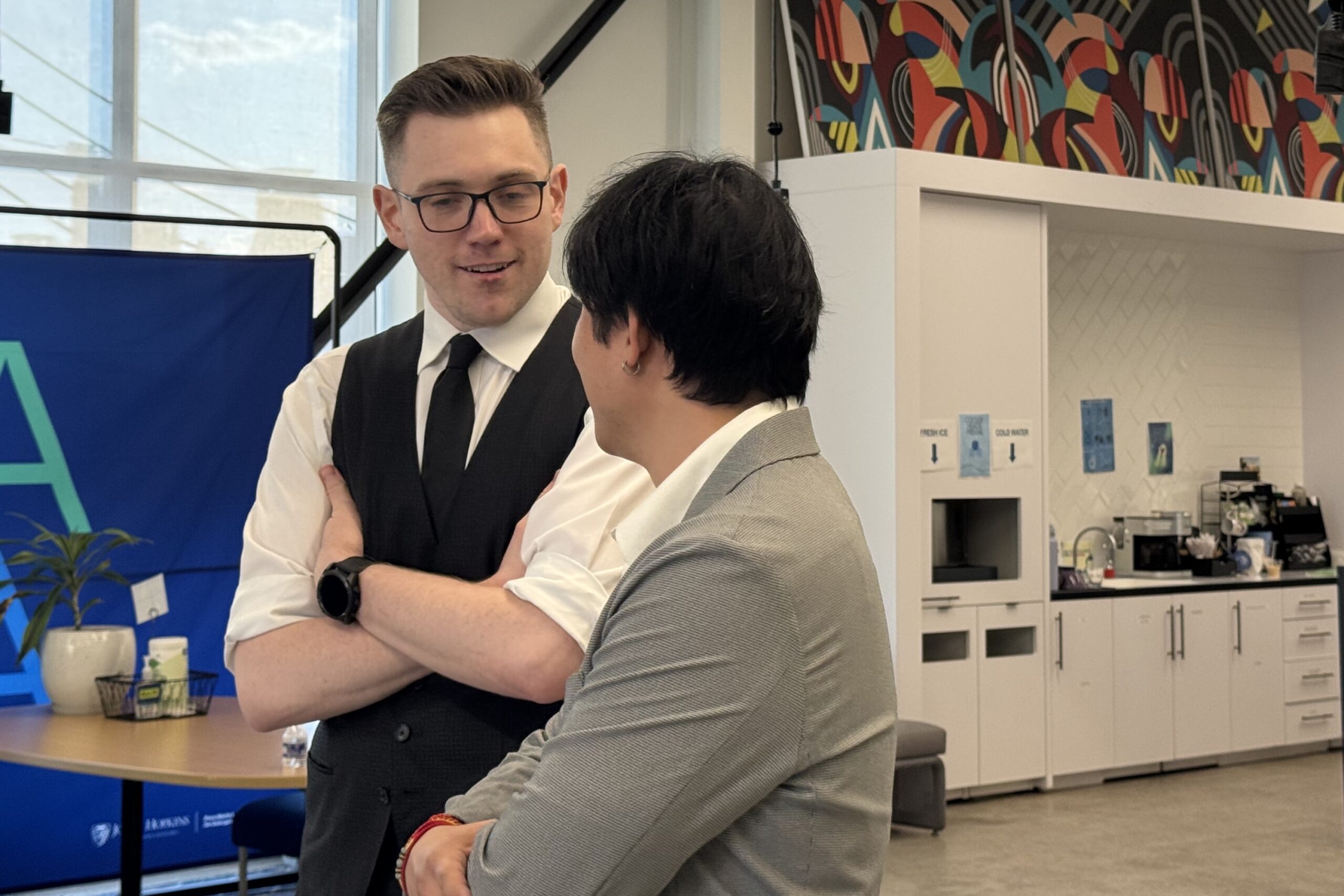 Two people in business attire have a conversation in a modern office space with a coffee area and colorful wall art in the background.
