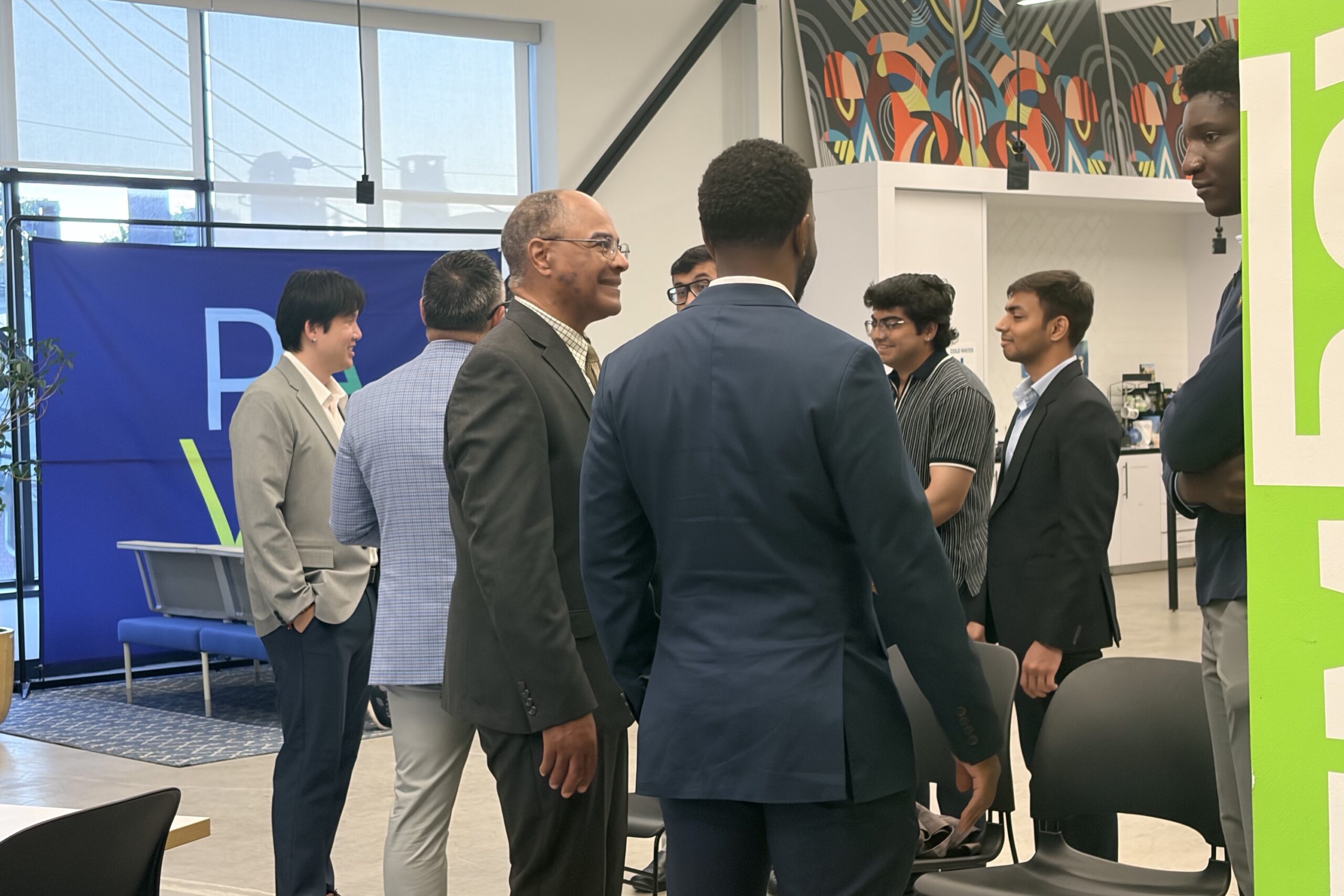 A group of people in business attire stand and talk in a modern office space with large windows and colorful wall art.