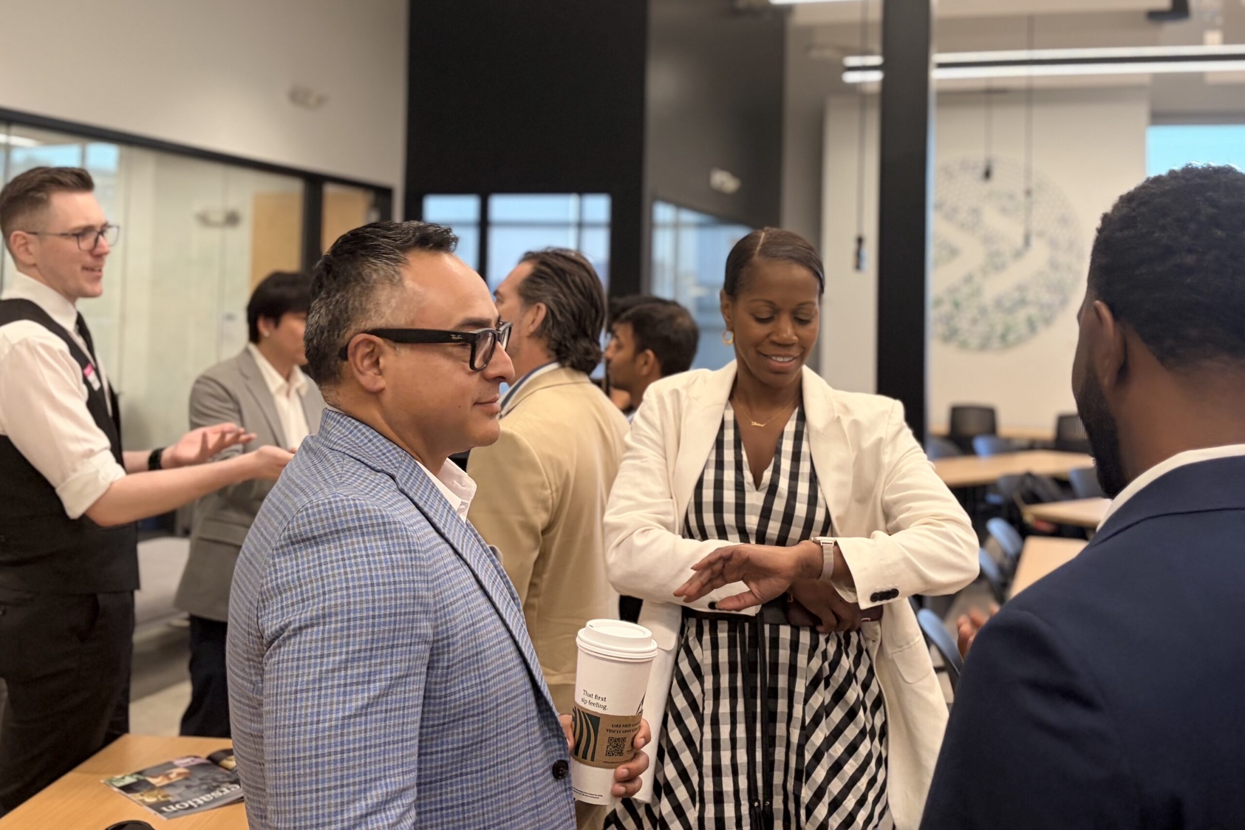 Several professionals stand and converse in a modern office meeting room; one woman checks her watch while a man nearby holds a coffee cup.