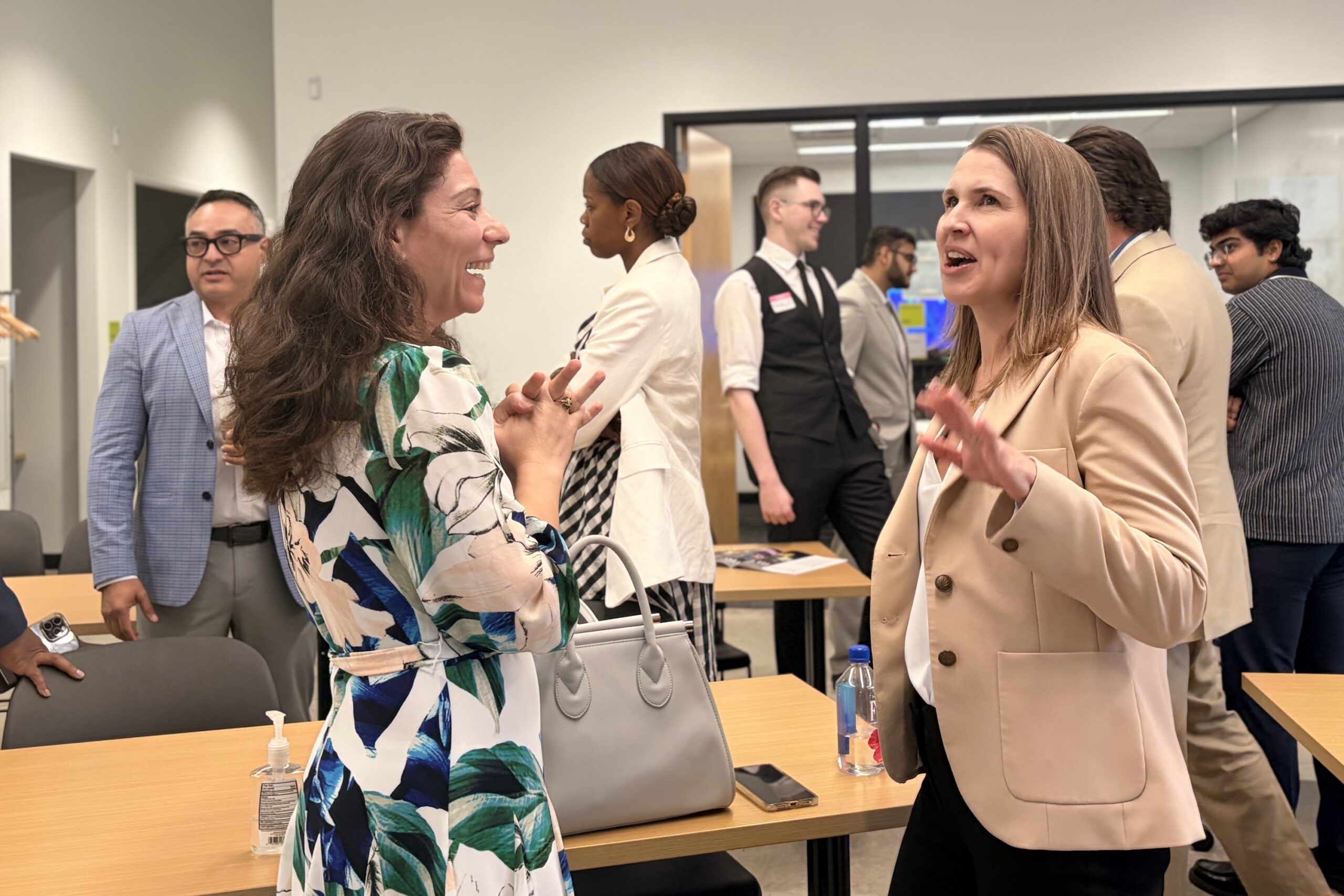 Two women stand and converse animatedly in a conference room as others talk in the background. Tables hold bags, a bottle, and hand sanitizer.