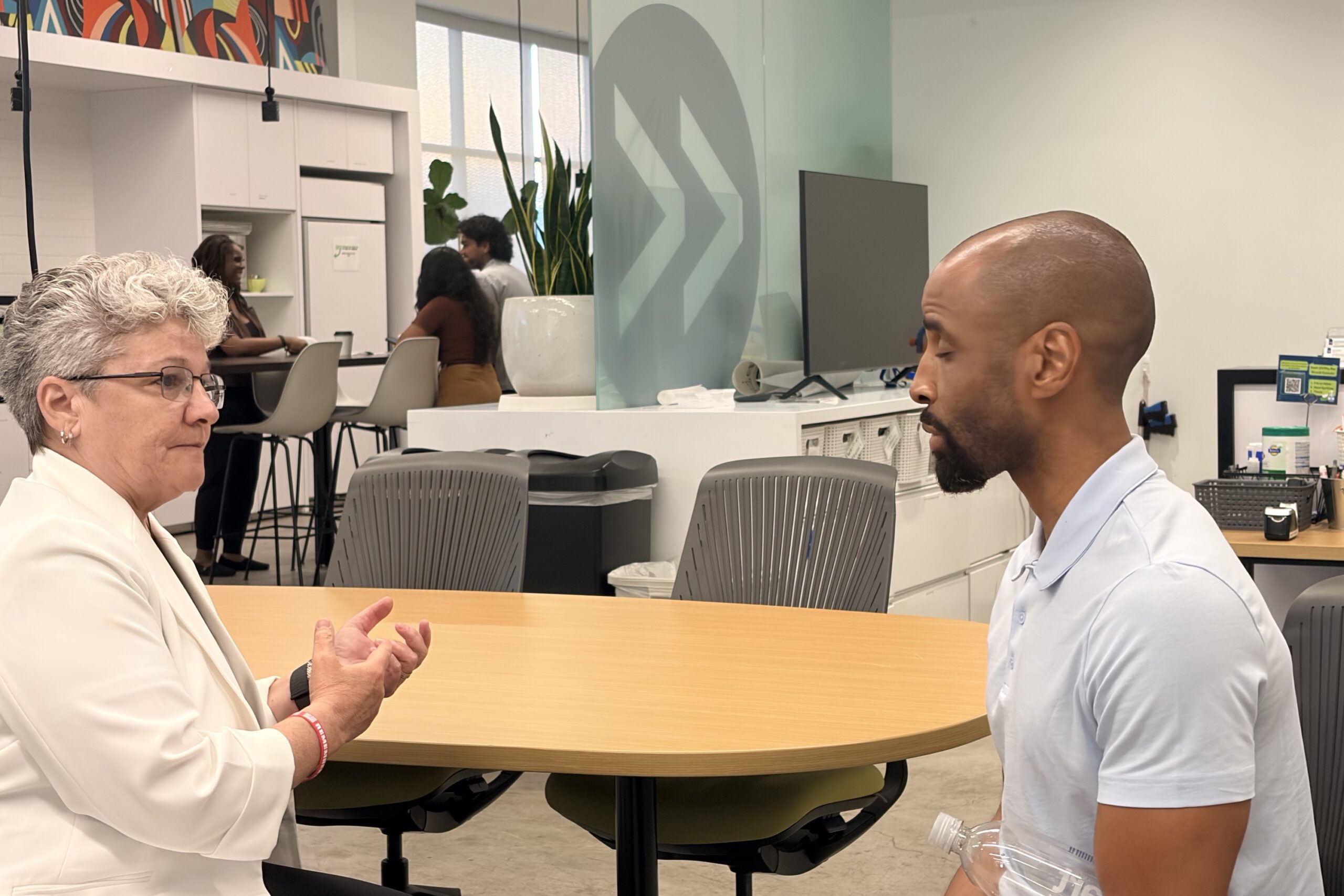 Two people sit across from each other at a round table in an office, engaged in conversation. Other people and office furniture are visible in the background.