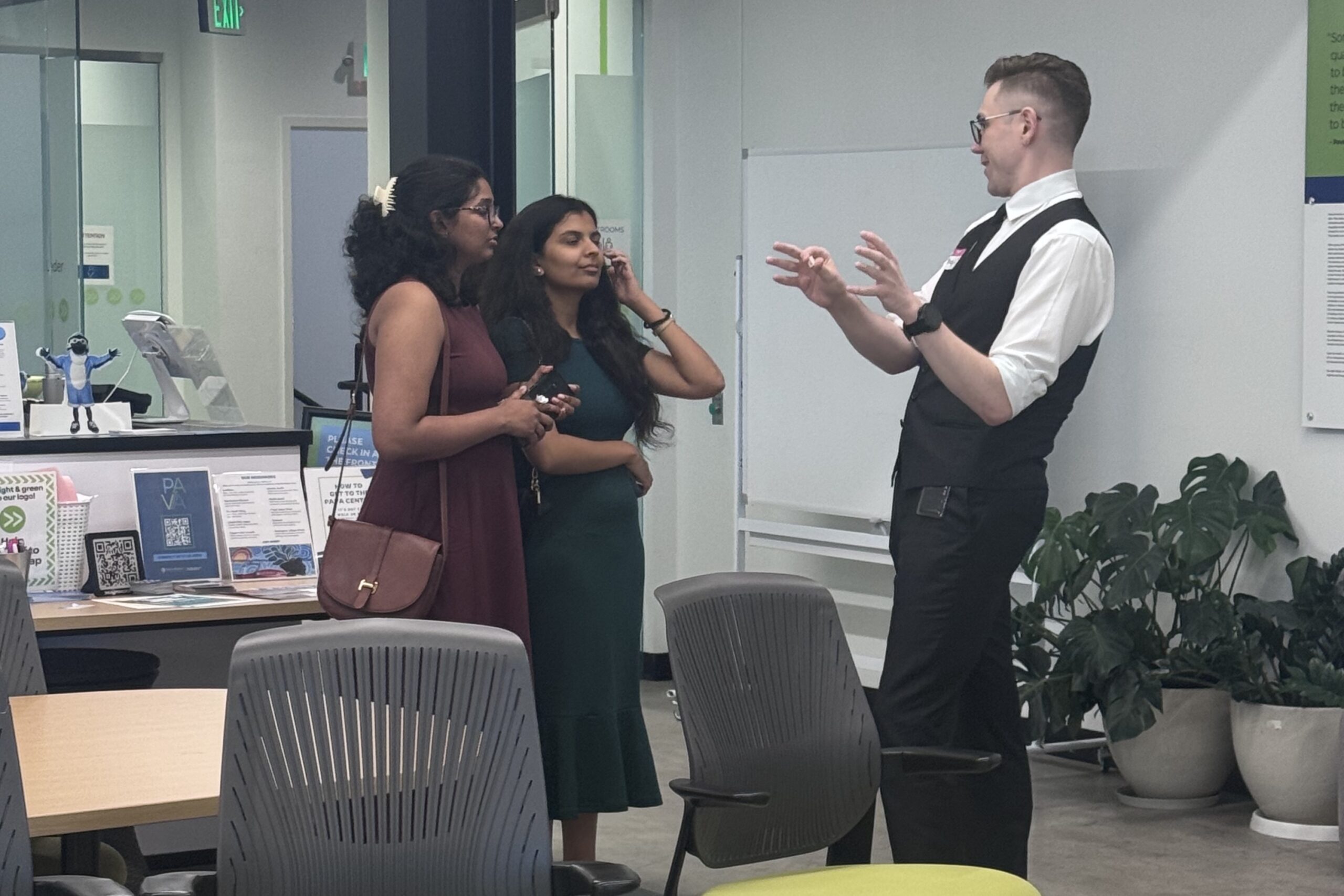 Three people stand and talk in an office or classroom space. Two women listen while a man in a vest gestures with his hands. Various chairs, plants, and informational signs are visible.
