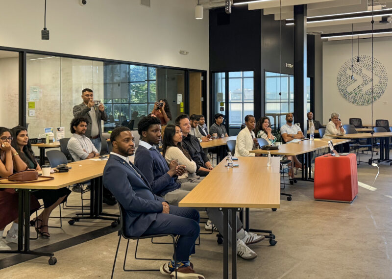 People are seated and listening attentively in a modern conference room, with a few individuals standing and taking photos.