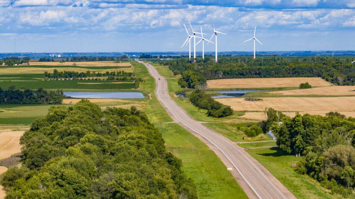 A rural road runs through fields and trees with several wind turbines in the distance under a partly cloudy sky.