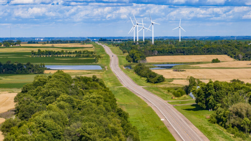 A rural road runs through fields and trees with several wind turbines in the distance under a partly cloudy sky.
