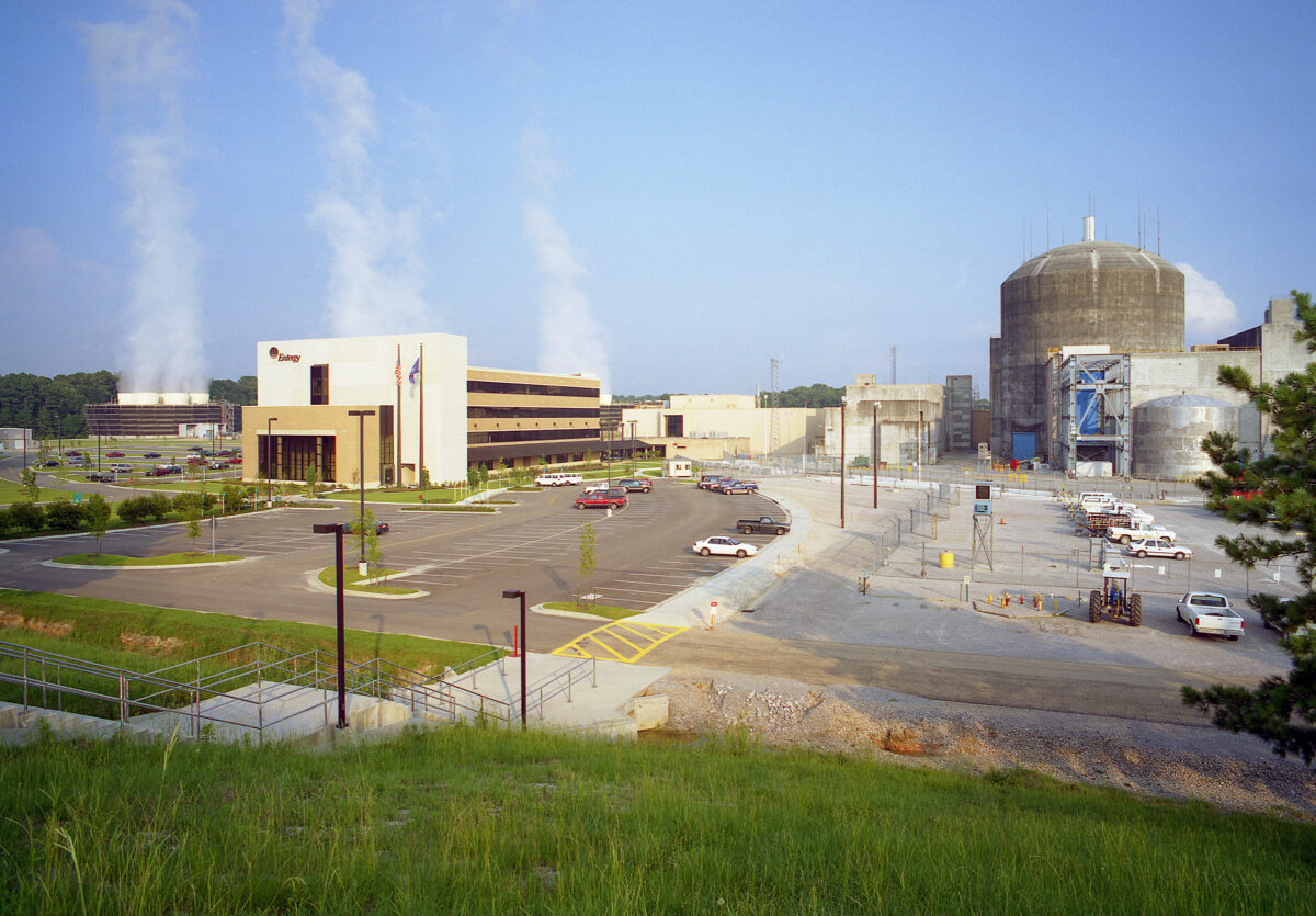 A nuclear power plant complex with cooling towers emitting steam, several buildings, parked cars, and a clear blue sky.