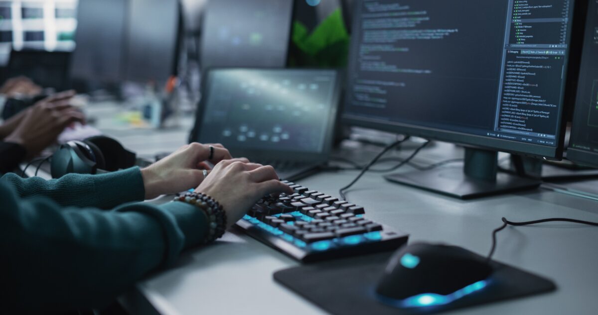 Hands typing on a keyboard in front of two monitors displaying code, with a mouse and headphones on the desk.