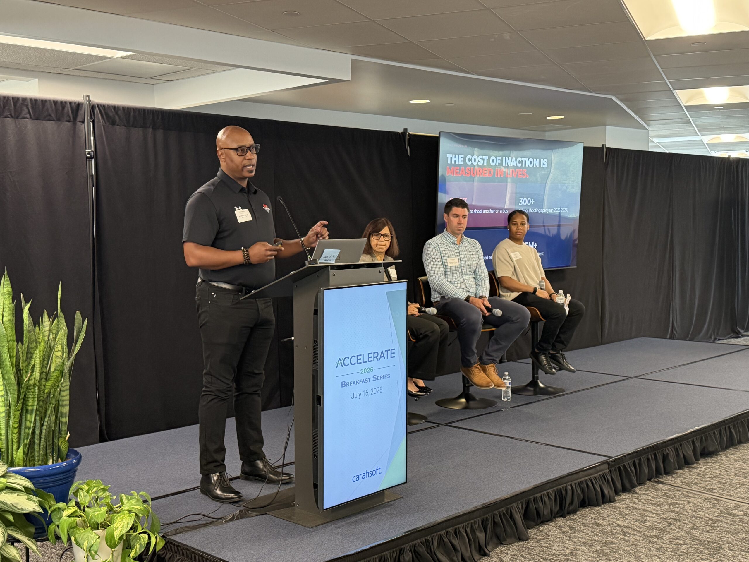 A man speaks at a podium labeled "ACCELERATE" while three seated panelists listen; a presentation slide is displayed in the background.
