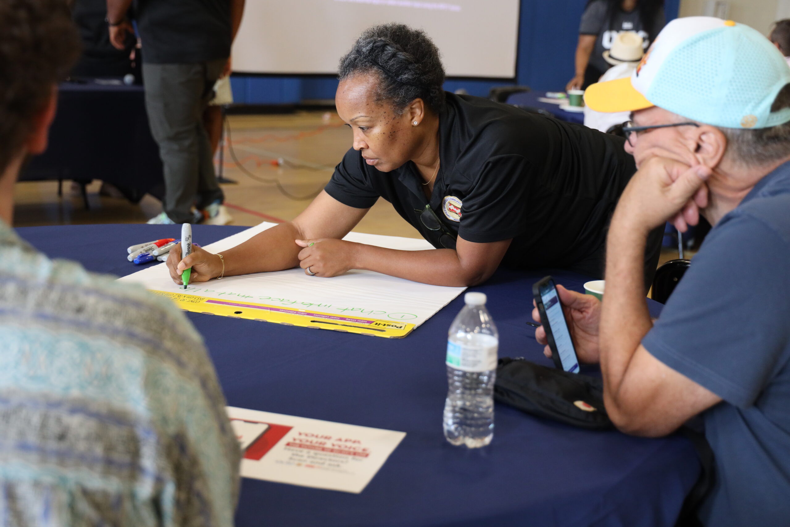 A woman leans over a table writing on large paper with a marker, while others sit nearby; a water bottle and phone are on the table.