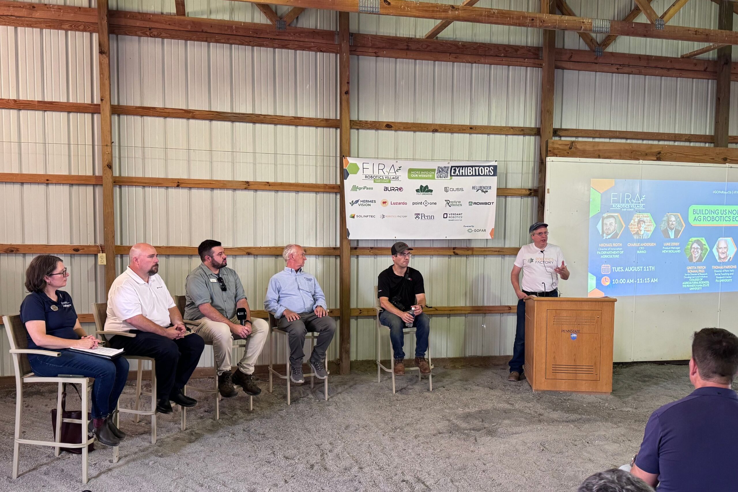 Six people sit or stand in front of a wooden wall and presentation screen at a panel event; one person is speaking at a podium while others listen.