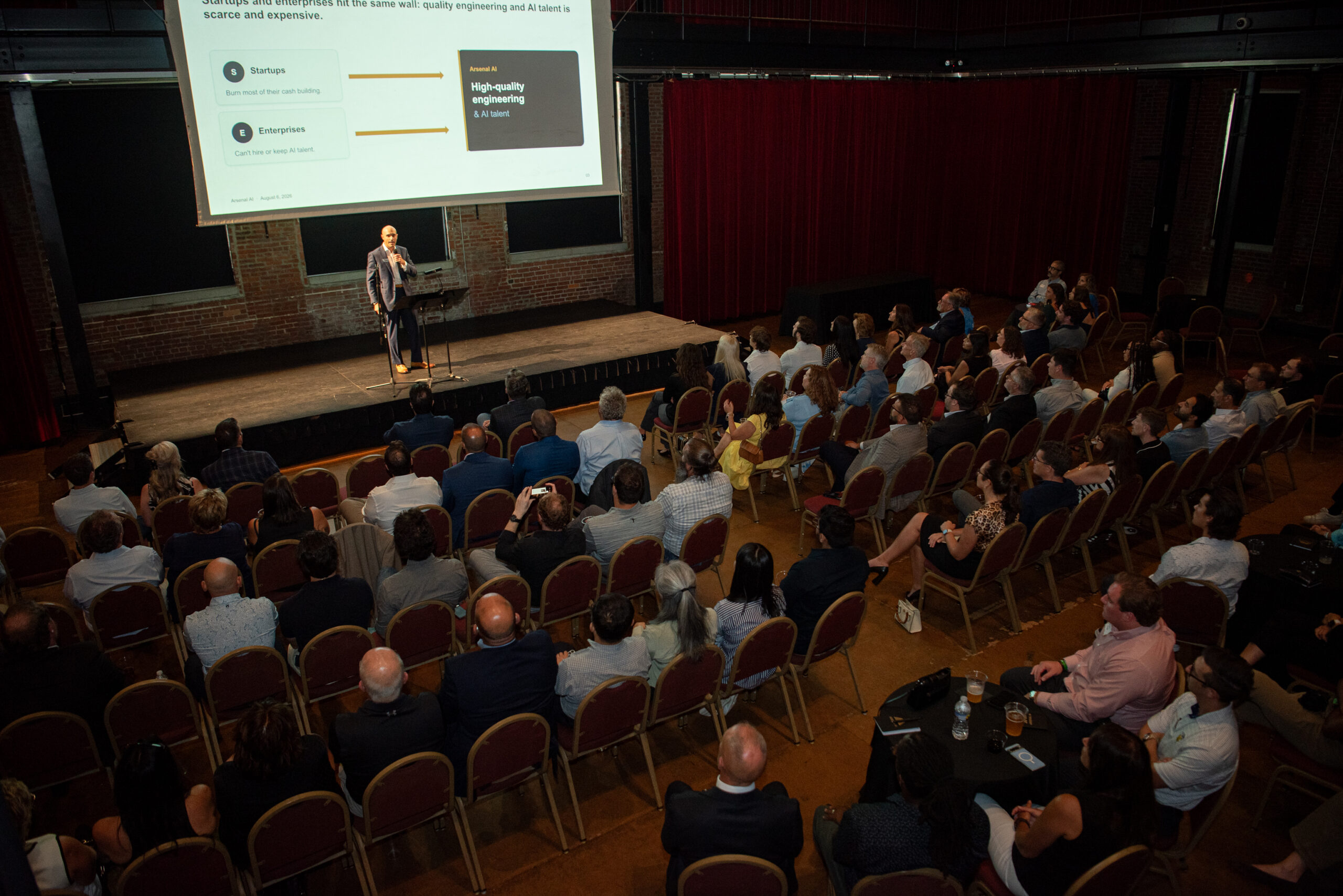 A speaker presents slides to an audience seated in rows at an indoor conference venue with red curtains and exposed brick walls.
