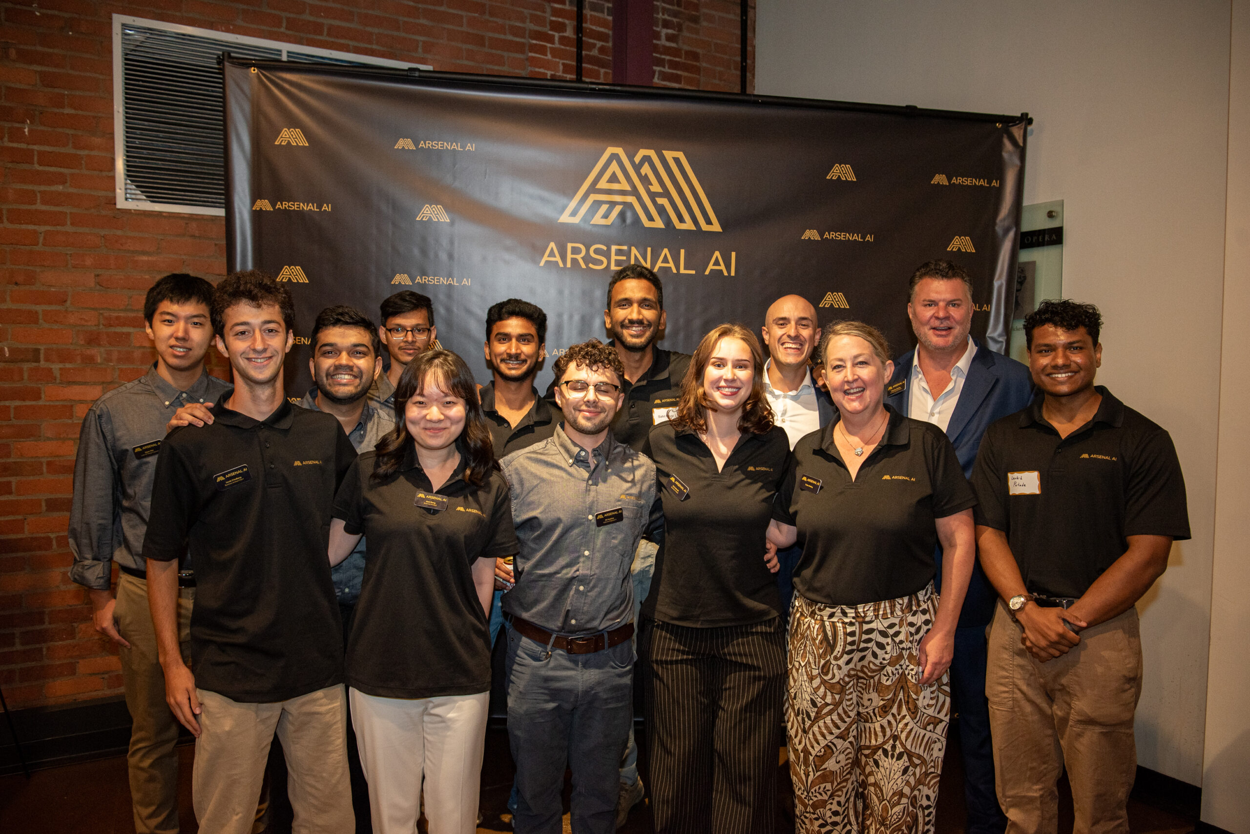 A group of thirteen people pose in front of an “Arsenal AI” banner at an indoor event, all smiling and casually dressed.