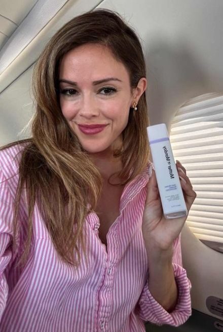 Woman in a pink striped shirt holds a white rectangular bottle, smiling, seated by an airplane window with sunlight coming through the shade.