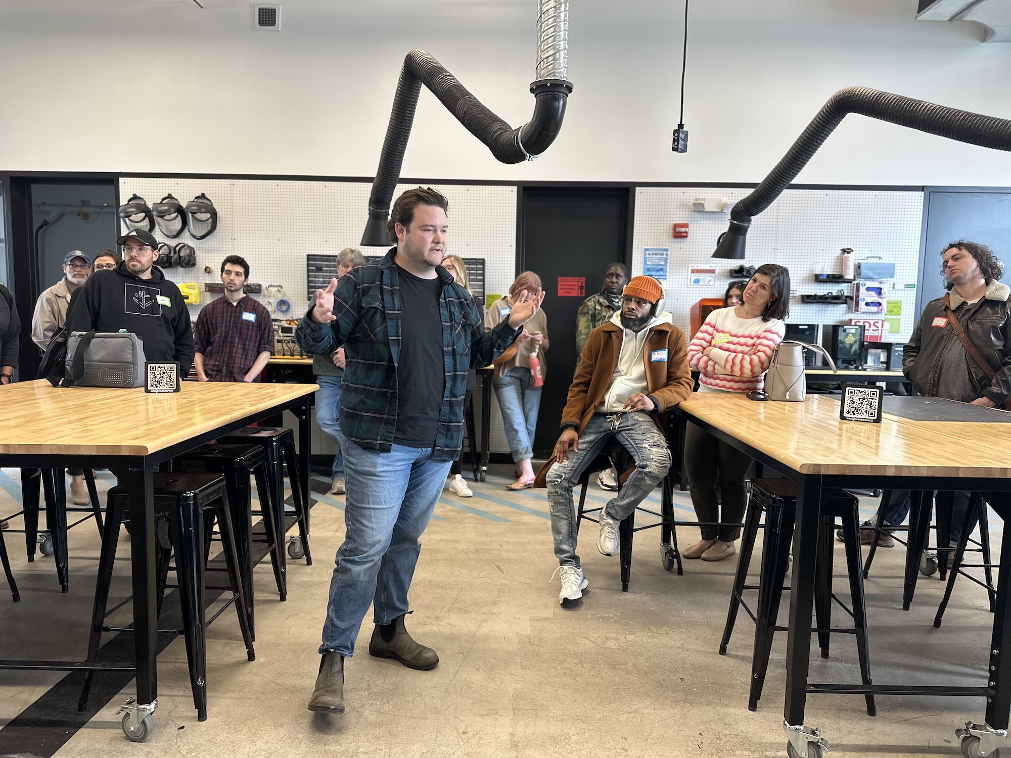 A man stands and gestures while speaking to a group gathered around tables in a modern workshop or classroom setting.