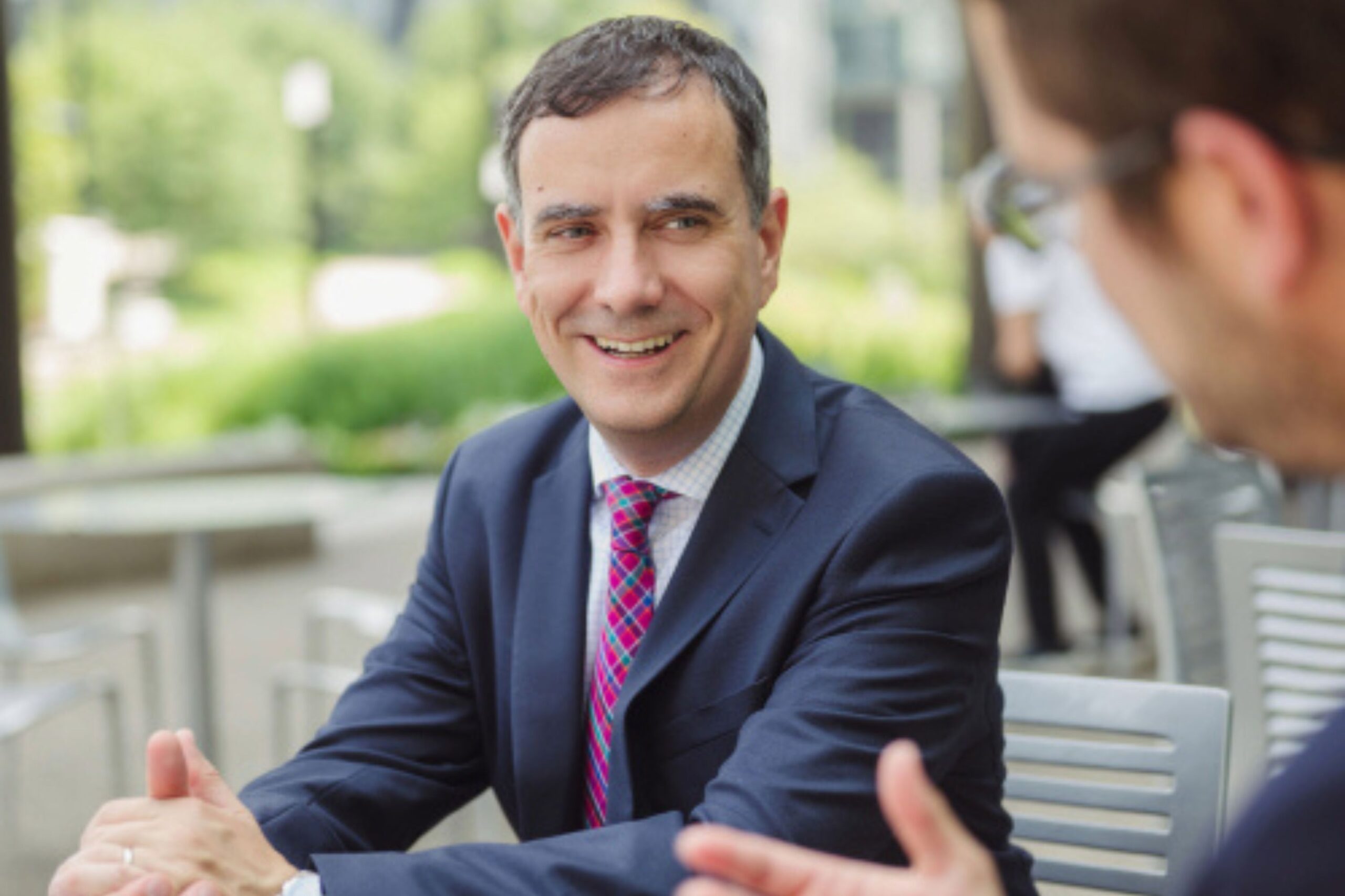 A man in a suit and tie sits outside at a table, smiling and engaged in conversation with another person.