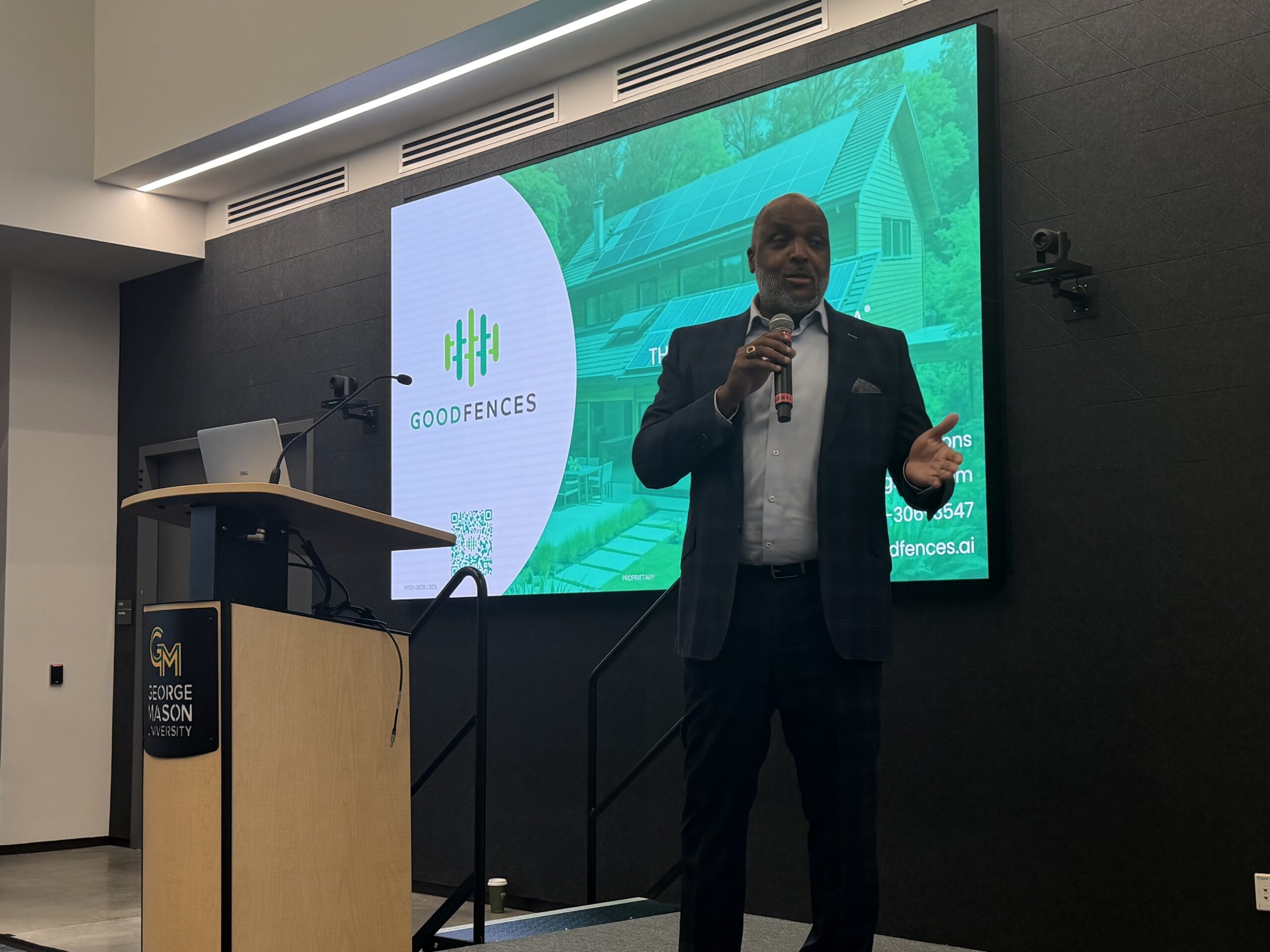 A man in a suit speaks on stage with a microphone at George Mason University. A screen behind him displays the GoodFences logo and a house image.