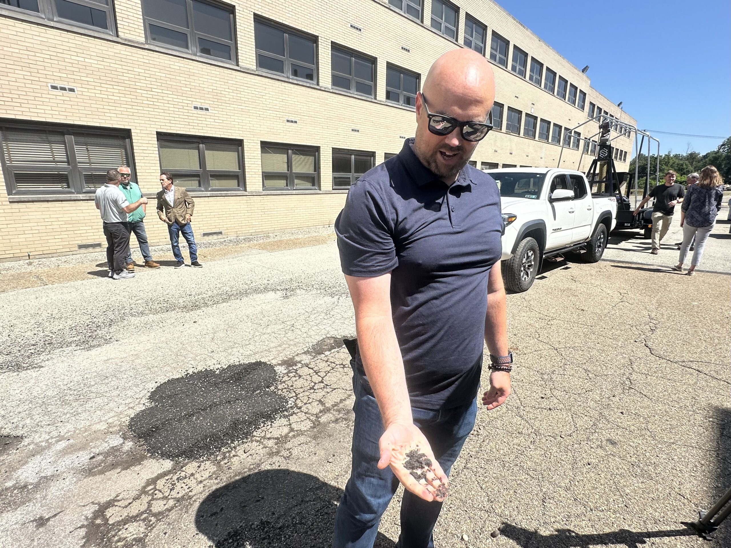 A man stands on a cracked pavement holding small rocks in his hand, with vehicles and people visible in the background near a large brick building.