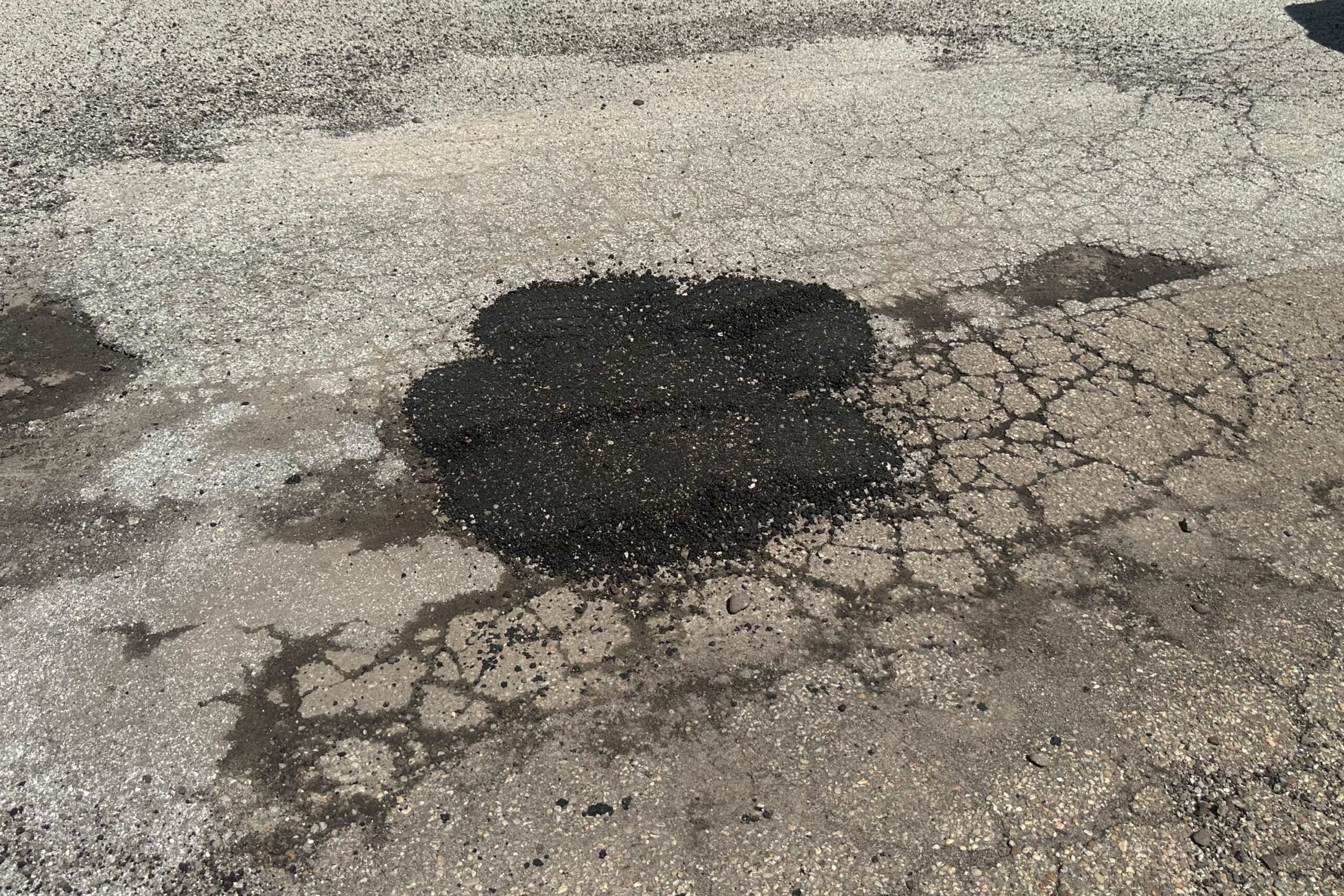 A patch of black asphalt covers a pothole on a cracked, weathered road surface.