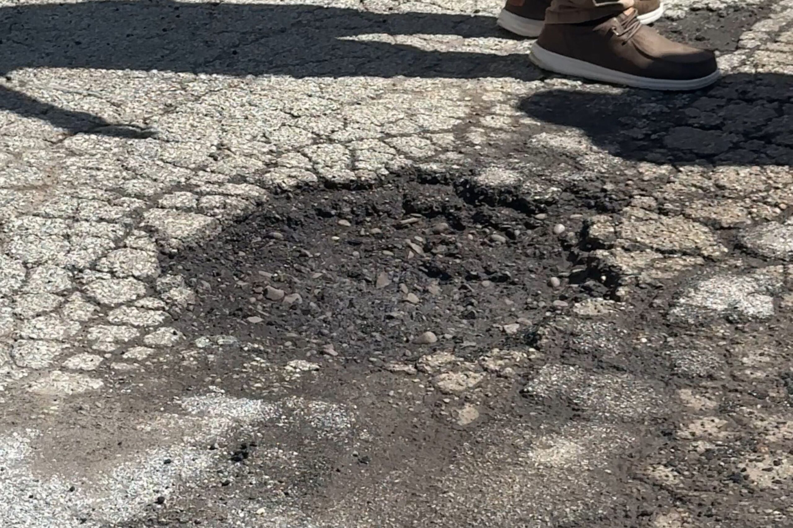 A large pothole in the middle of a cracked asphalt road with a person’s feet visible nearby.