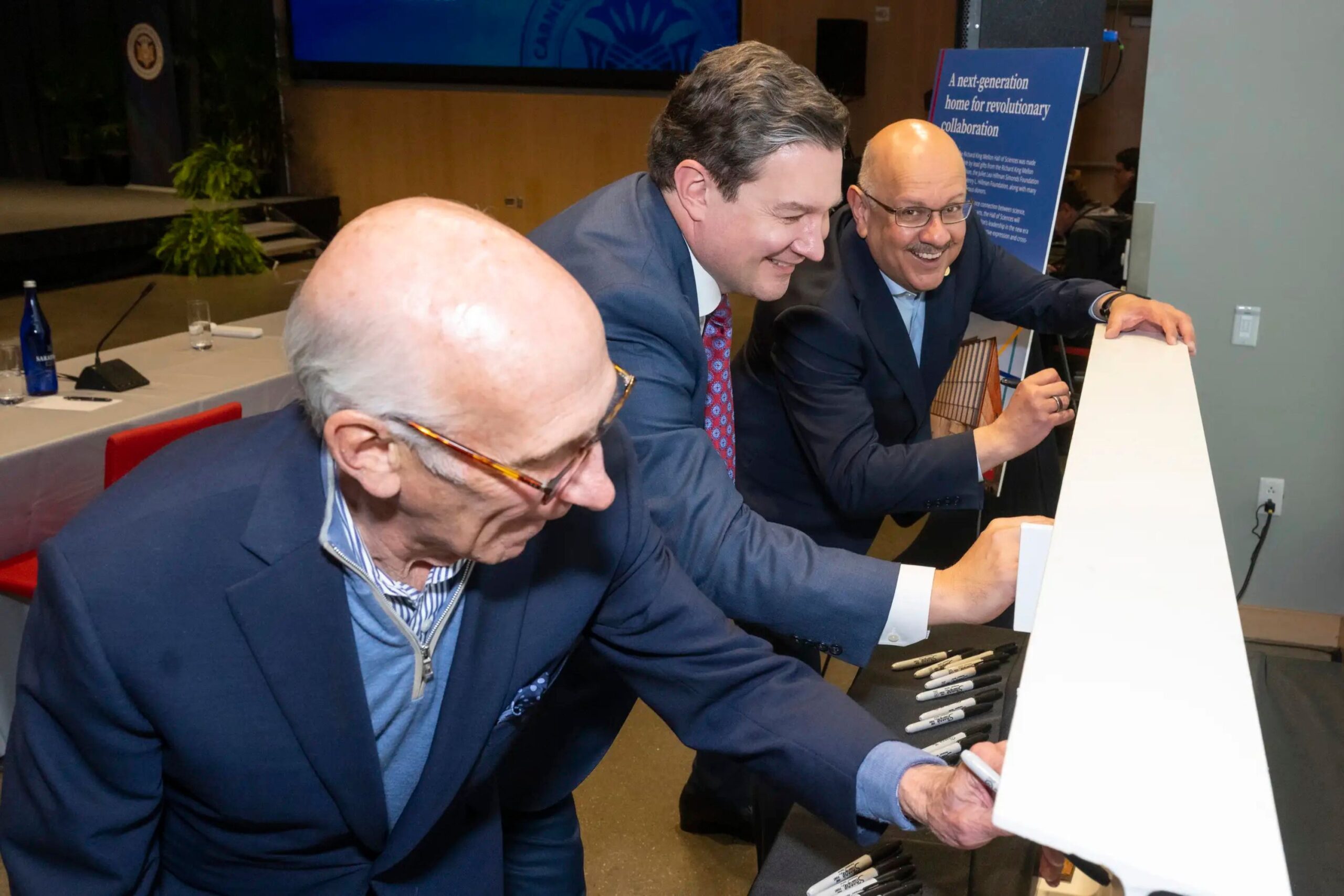 Three men in suits sign a white beam with markers at an indoor event, with a table, chairs, and informational poster visible in the background.