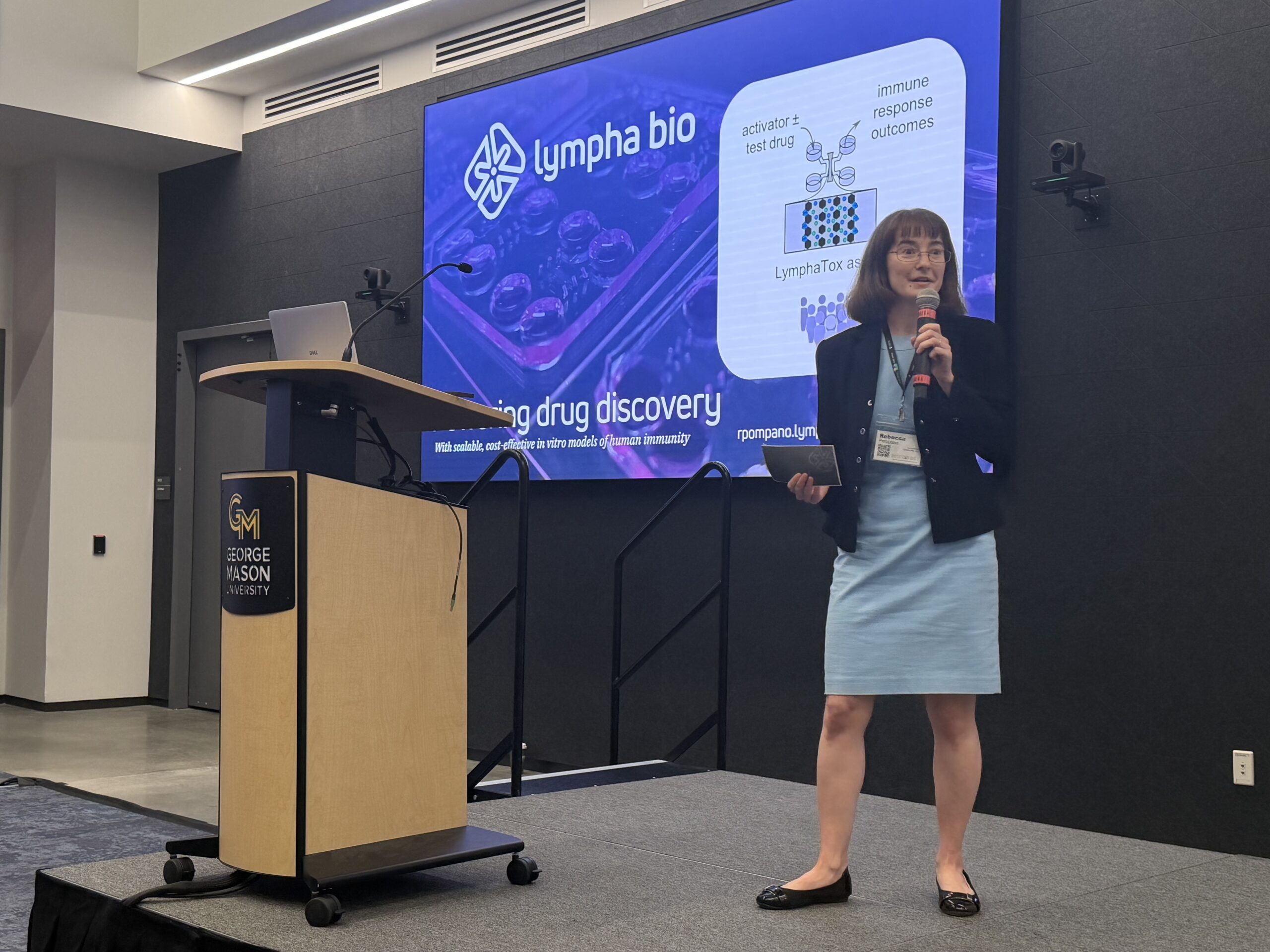A woman stands on stage giving a presentation on drug discovery for Lympha bio at George Mason University, with a slideshow projected behind her.