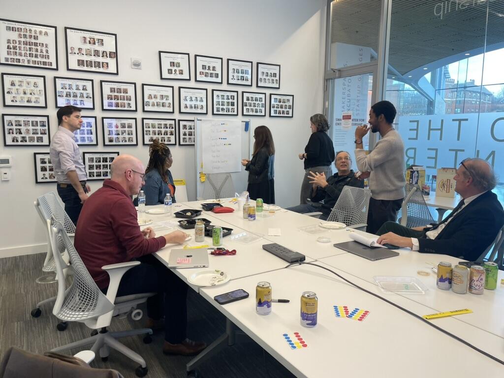 Seven people in a modern conference room have a meeting with whiteboards and laptops. Some are seated at a table with drinks and snacks, while others stand and discuss near the board.