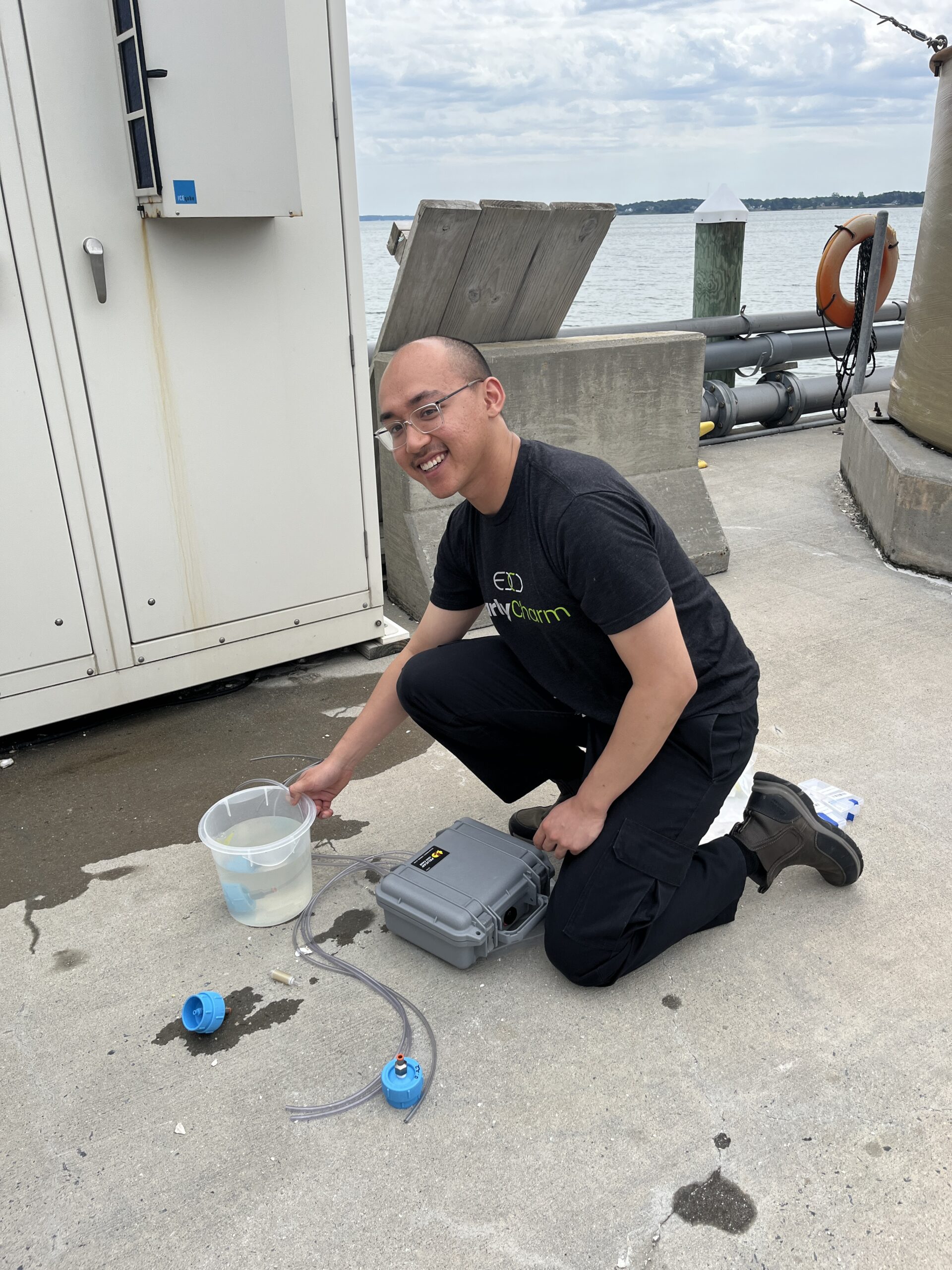 A person kneels on a concrete dock next to scientific equipment, holding a clear container of water with a hose, near the edge of a body of water under a cloudy sky.