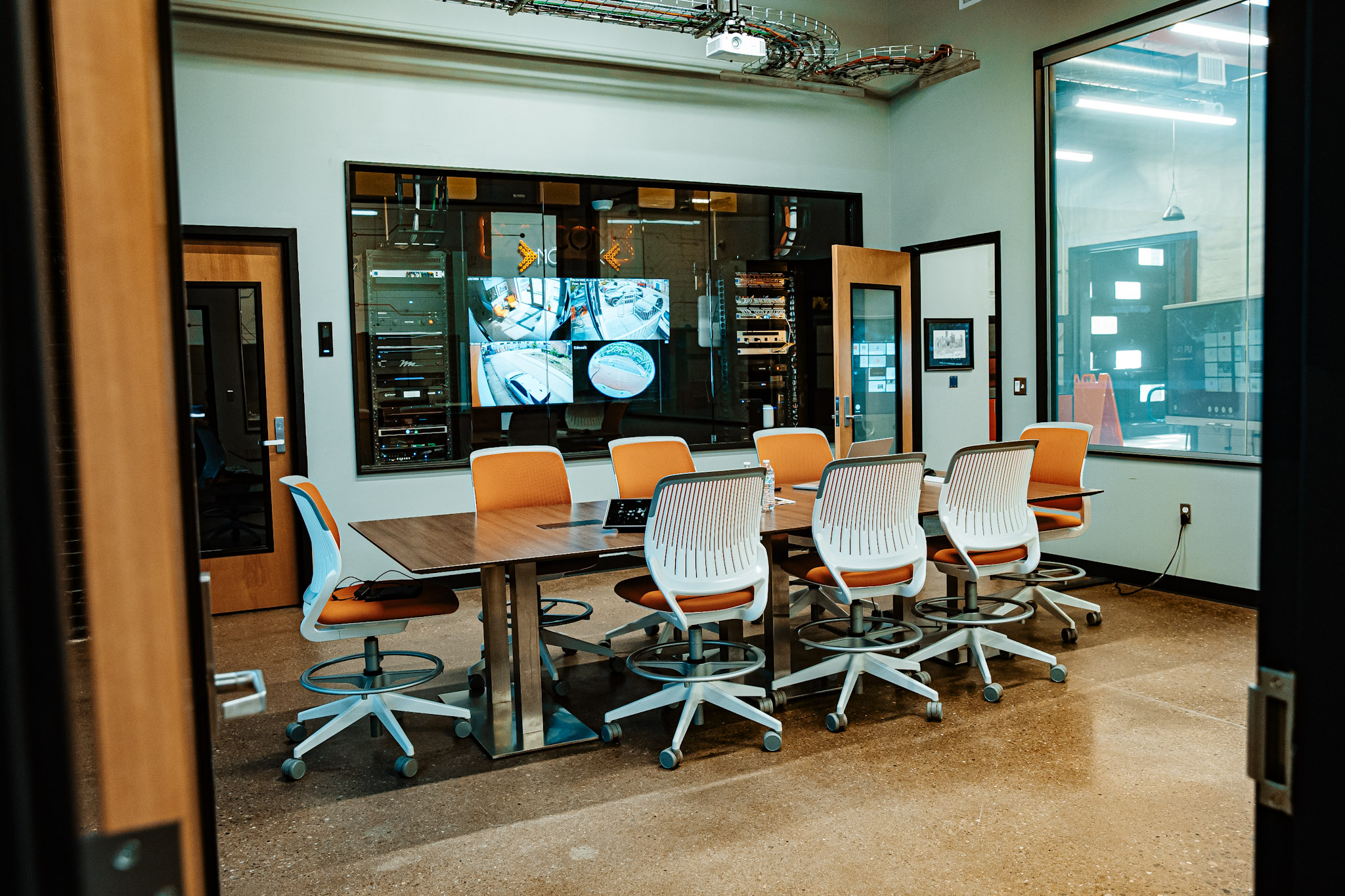 Conference room with an empty table surrounded by orange and white chairs, large wall window, visible security monitors, and high-tech equipment in the background.