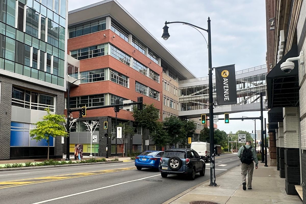 A city street with modern buildings, a skywalk, traffic lights, cars, pedestrians, and a black banner reading “AVENUE.”.