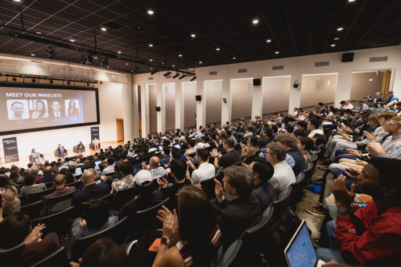 A large audience sits in an auditorium facing a stage with a panel discussion. A screen displays “Meet Our Panelists” with photos and names of four speakers.