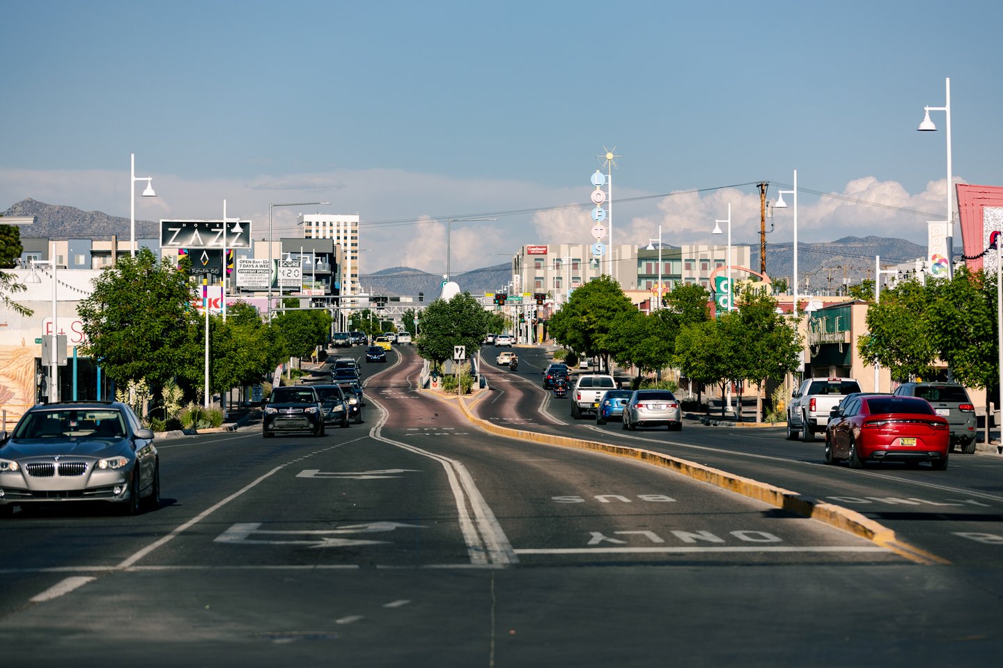 A city street with cars driving in both directions, lined with trees, buildings, and traffic signs under a clear blue sky.