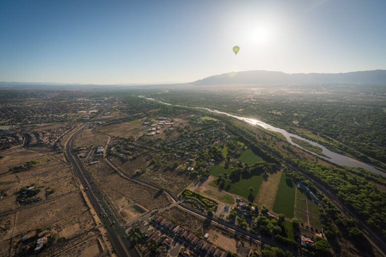 Aerial view of a cityscape with a river, green fields, and residential areas under a clear sky; a hot air balloon floats near the sun above distant mountains.