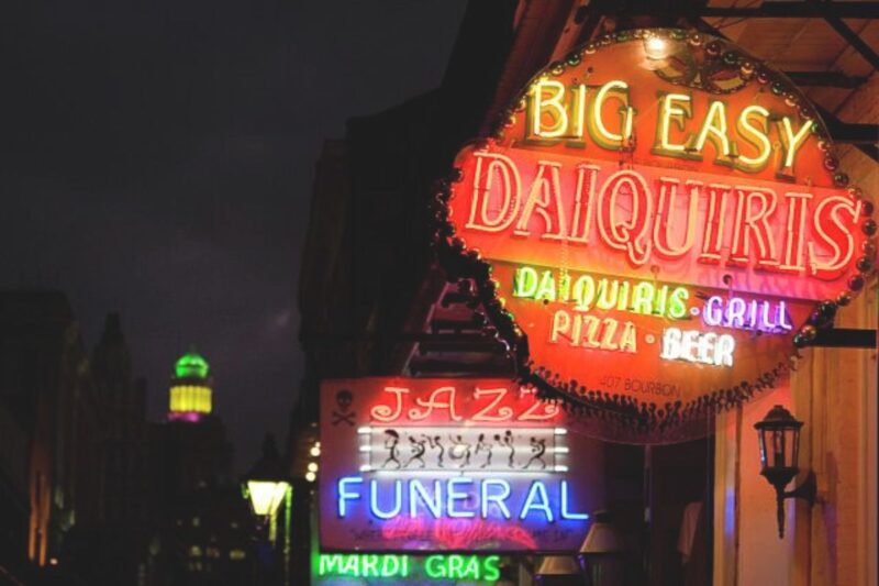 Neon signs on a street at night advertise “Big Easy Daiquiris,” a jazz funeral, and Mardi Gras, with buildings and a glowing green tower in the background.