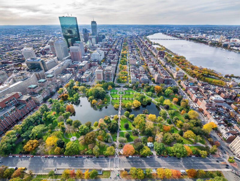 Aerial view of Boston Public Garden with surrounding city buildings, tree-lined streets, and the Charles River visible in the background.