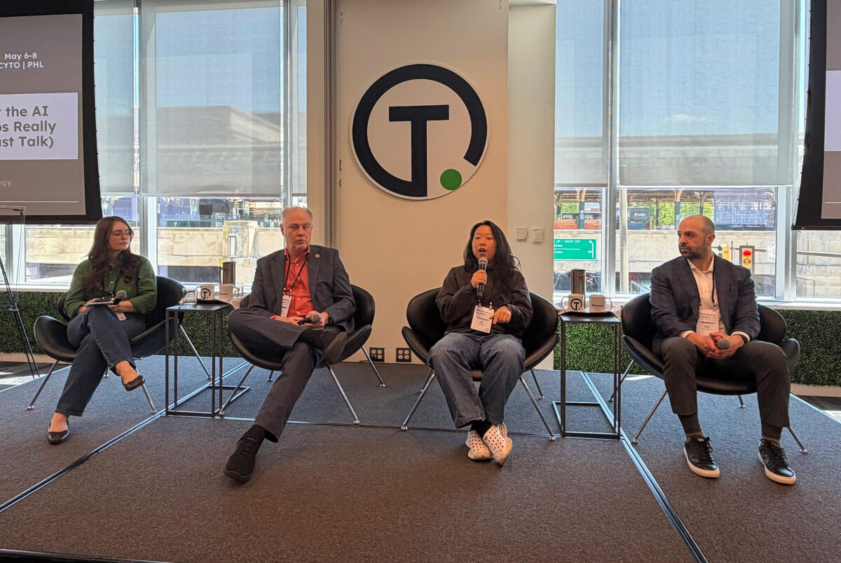 Four people sit on a panel on stage in front of large windows and a circular logo with a “T” in the center. One panelist holds a microphone and speaks while the others listen.