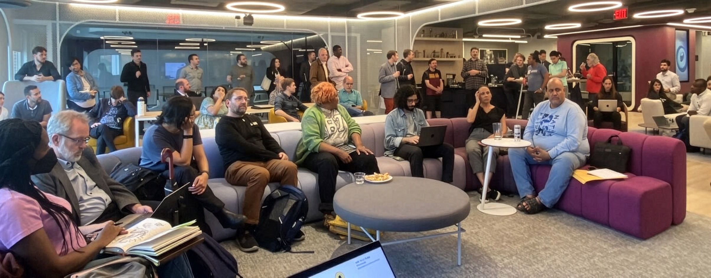 A diverse group of people sit and stand in a modern lounge, listening to a presentation. Some have notebooks or laptops, and refreshments are on tables.