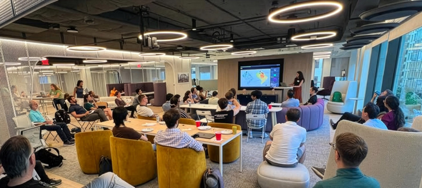 A group of people sits in a modern office space, watching a presentation projected on a screen at the front of the room.