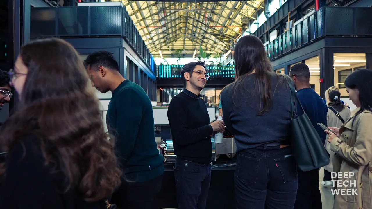 People stand and talk in a modern, high-ceilinged building during an event. “DEEP TECH WEEK” is visible in the bottom right corner.