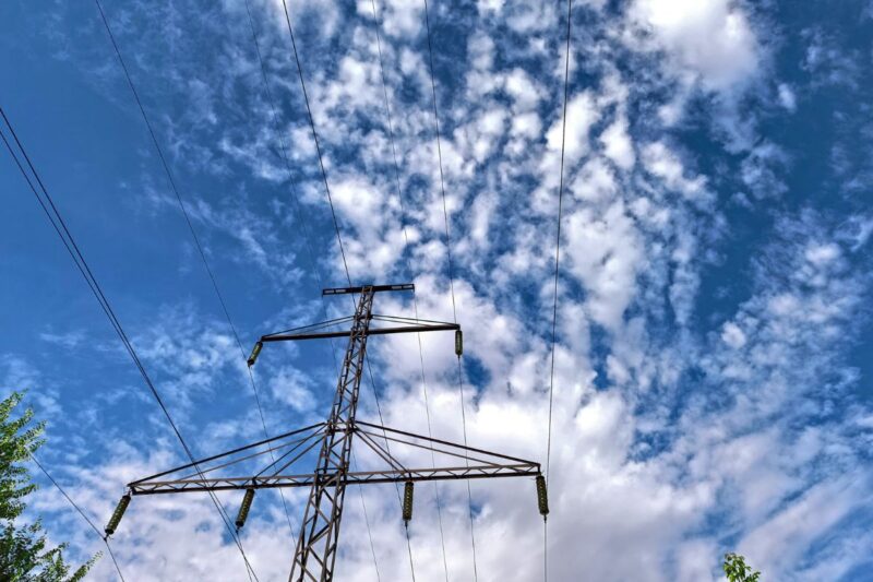 A metal electricity transmission tower with multiple power lines stretches upward against a blue sky with scattered white clouds.