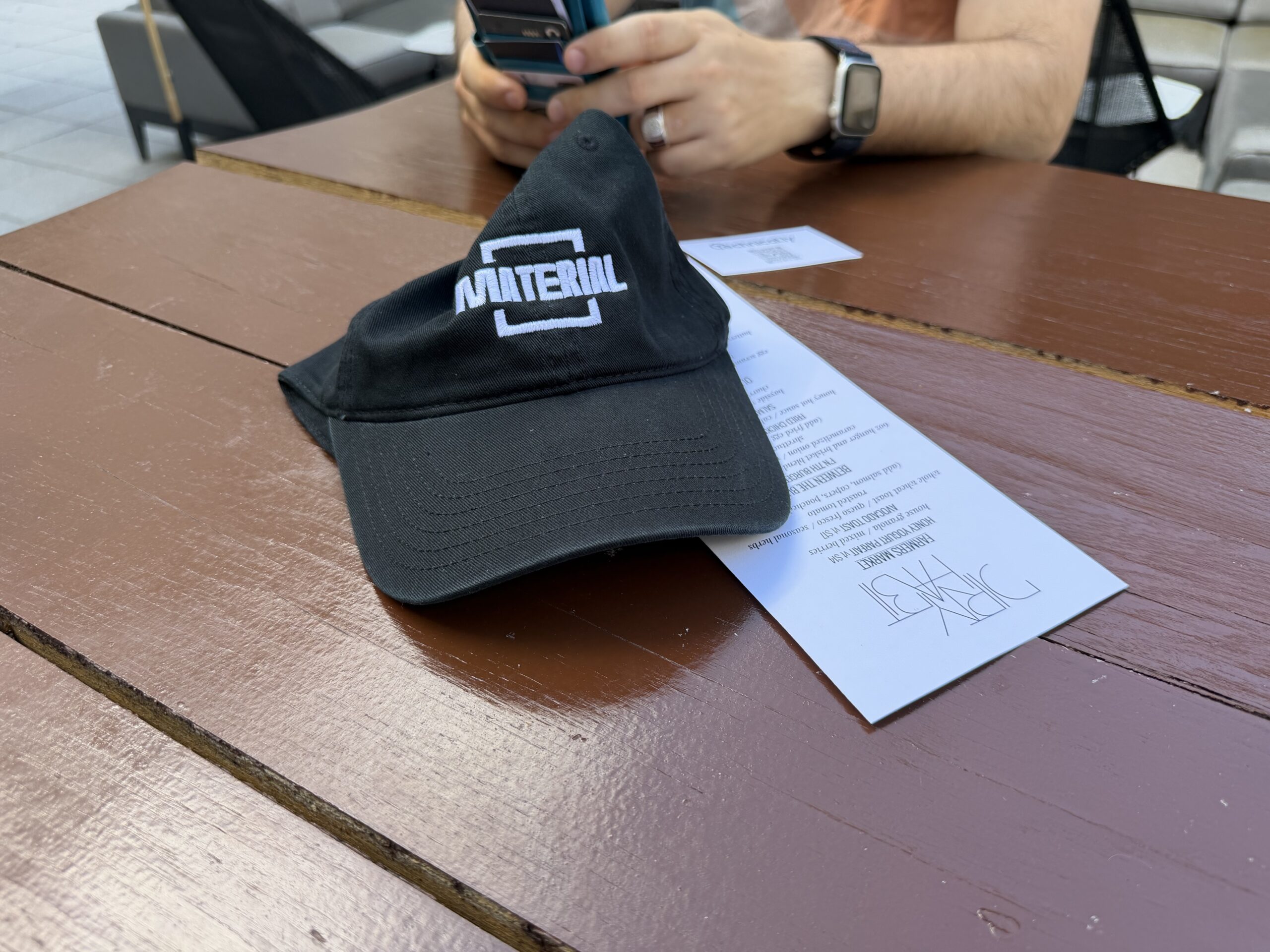 A black cap with “MATERIAL” text rests on a brown wooden table next to a menu. In the background, a person uses a smartphone.