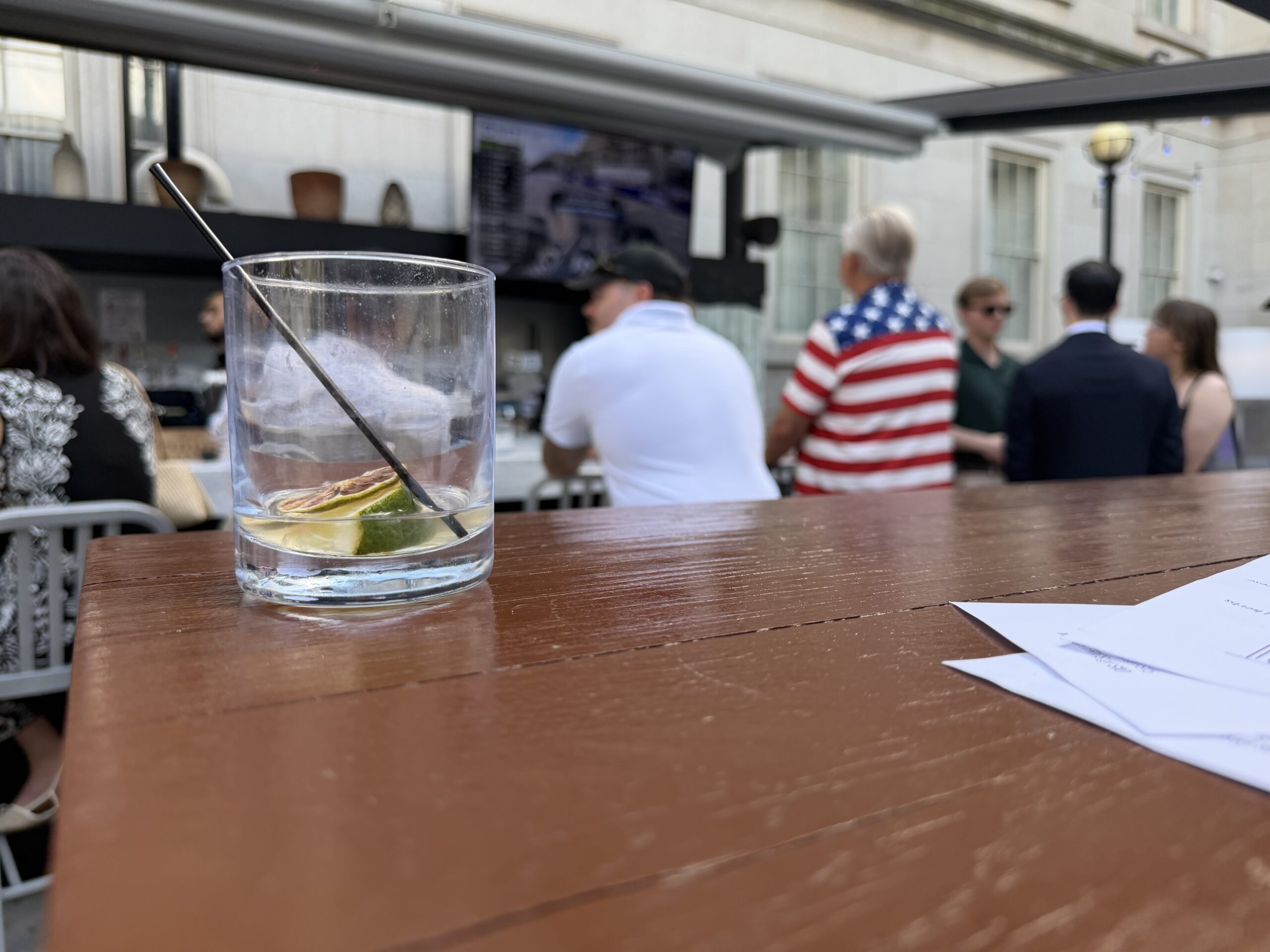 A glass with lime and a straw sits on a wooden table; people stand and sit at an outdoor bar in the background.